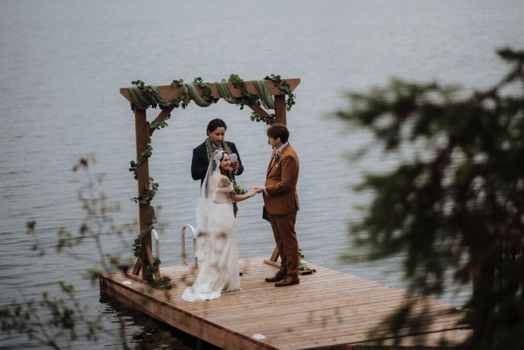A couple on a dock on a moody lake at Falcon Trails Resort for their wedding. They are holding hands, and dressed in their wedding clothes during their wedding ceremony. The officiant stands behind them under their arch.
