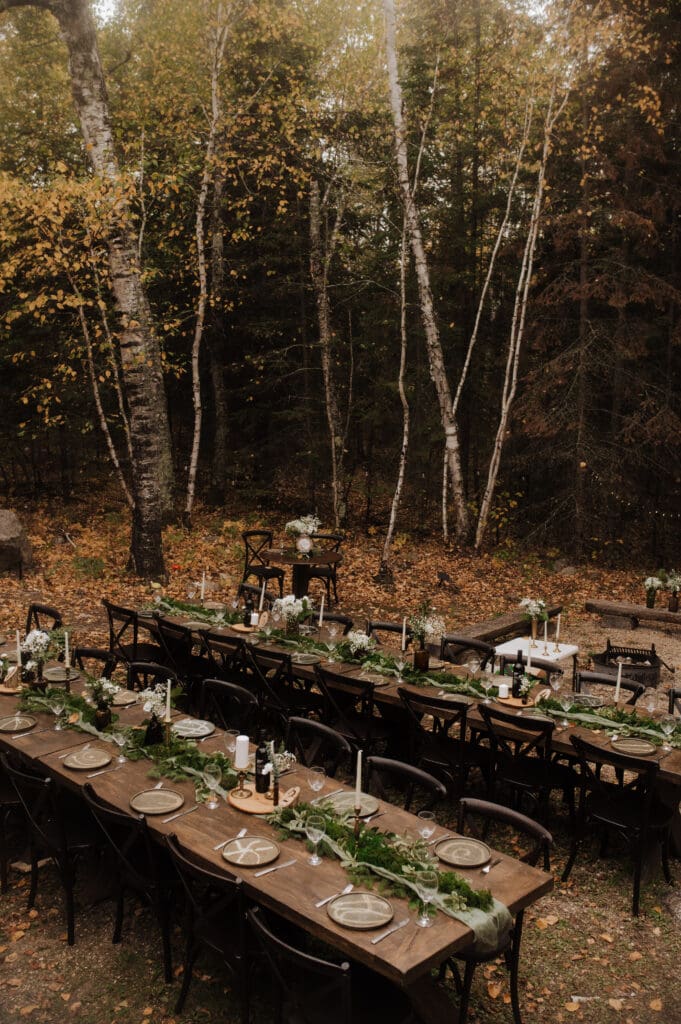 A scene in a forest right out behind a cabin at Falcon Trails Resort. Two harvest tables are decorated with greenery and flowers, acting as a reception area. There are candles and bottles of wine on the table, and the photo is taken before the reception has happened, so everything looks perfect.