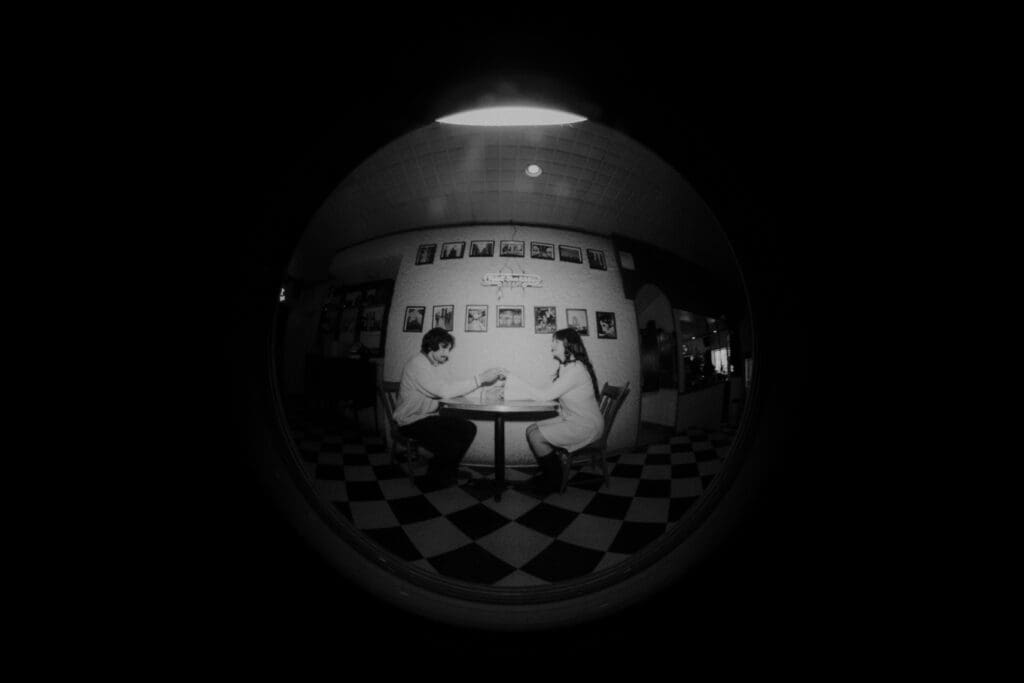 A black and white photo of a couple during their engagement session at Winnipeg's favourite local pizza joint, Shorty's Pizza. The image is shot with a fish eye lens to give it a security camera look.