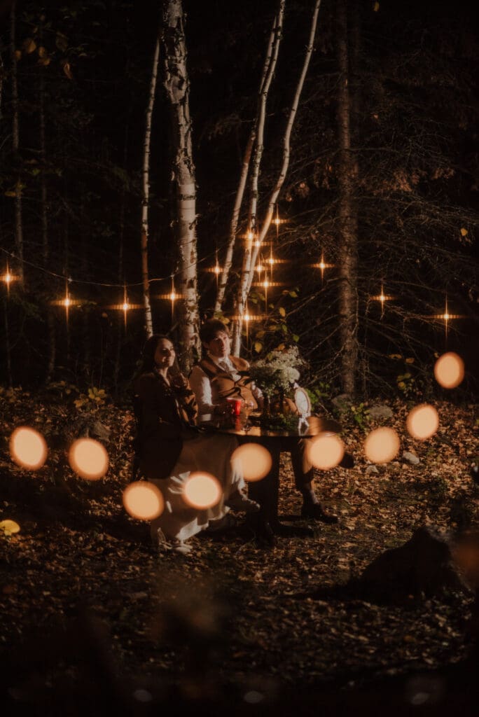 The married couple sits underneath twinkly lights in their reception area at Falcon Trails Resort, listening to speeches, and eating dinner. They are wearing theie wedding clothes and the bride is wearing a coat, as the sun has gone down. The lights are warm and romantic, and they sit in a clearing of trees behind the cabin.